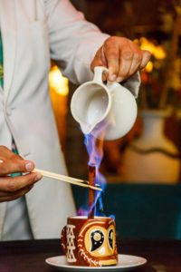 Purple flames rise during the creation of a Kona coffee in a bold tiki mug.
